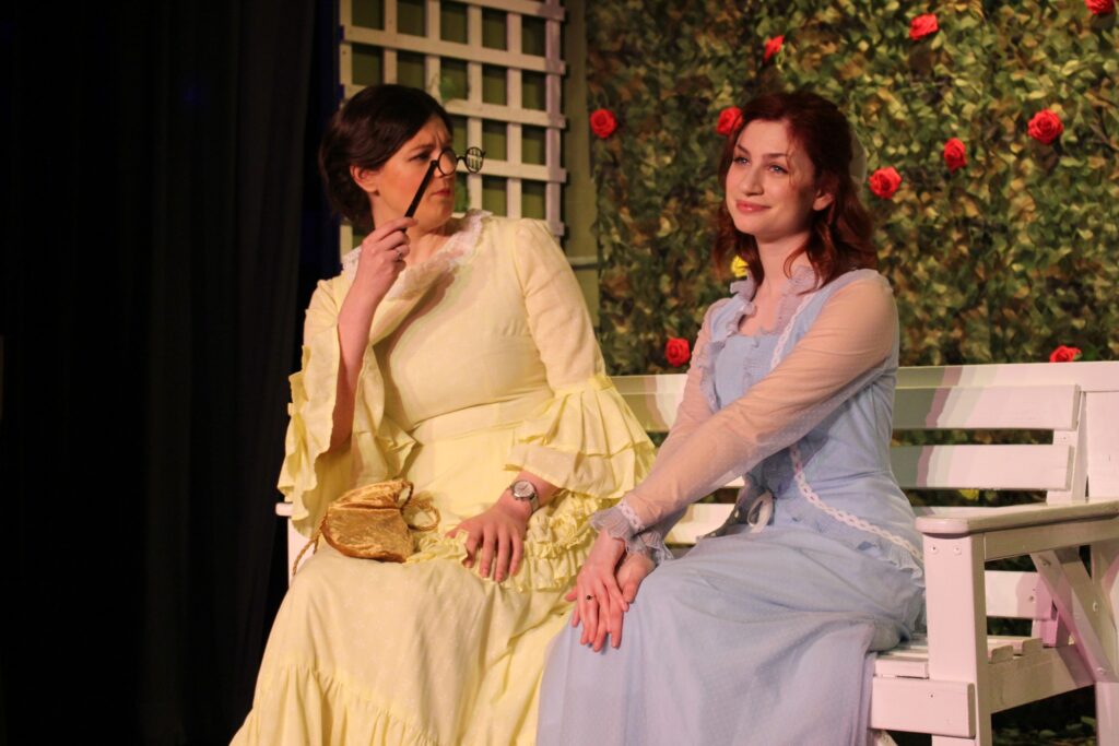 Gwendolen Fairfax observes Cecily Cardew appraisingly through her glasses. Both young women are seated side by side on a white bench. Behind them, you can see a trellis and hanging ivy with dotted red roses. Gwendolen is dressed in a pastel yellow dress with draping sleeves. Cecily is posing to be observed, looking out to the audience with a vapid smile. Her dress is pastel blue, with sheer sleeves.