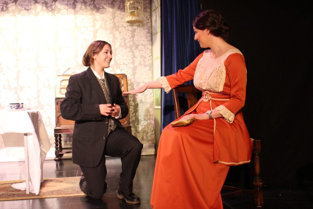 Gwendolen is sitting on a chair, dressed in a dark orange dress with cream lace over the bodice. She is holding out her hand imperiously to Jack, who is kneeling next to the chair. Jack is wearing a dark suit.